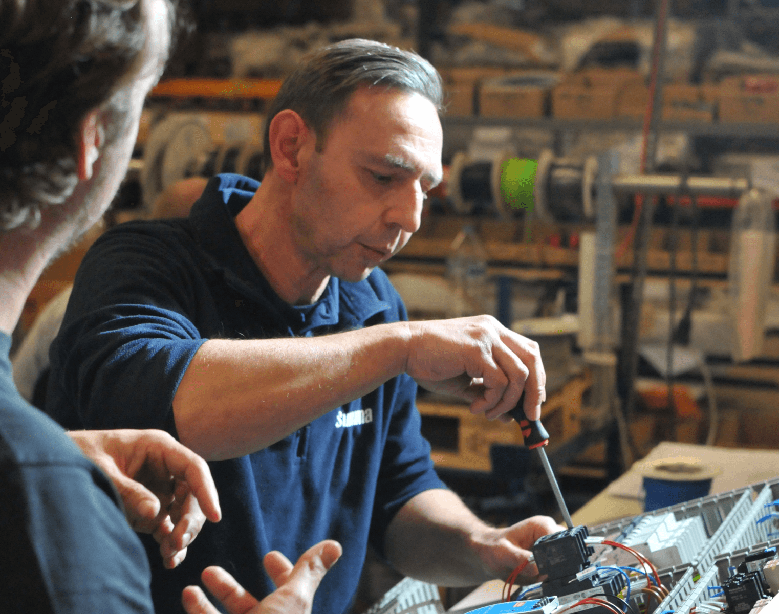 Two men collaborate on an electronic project in a workshop, focusing on a circuit board with a screwdriver in hand.
