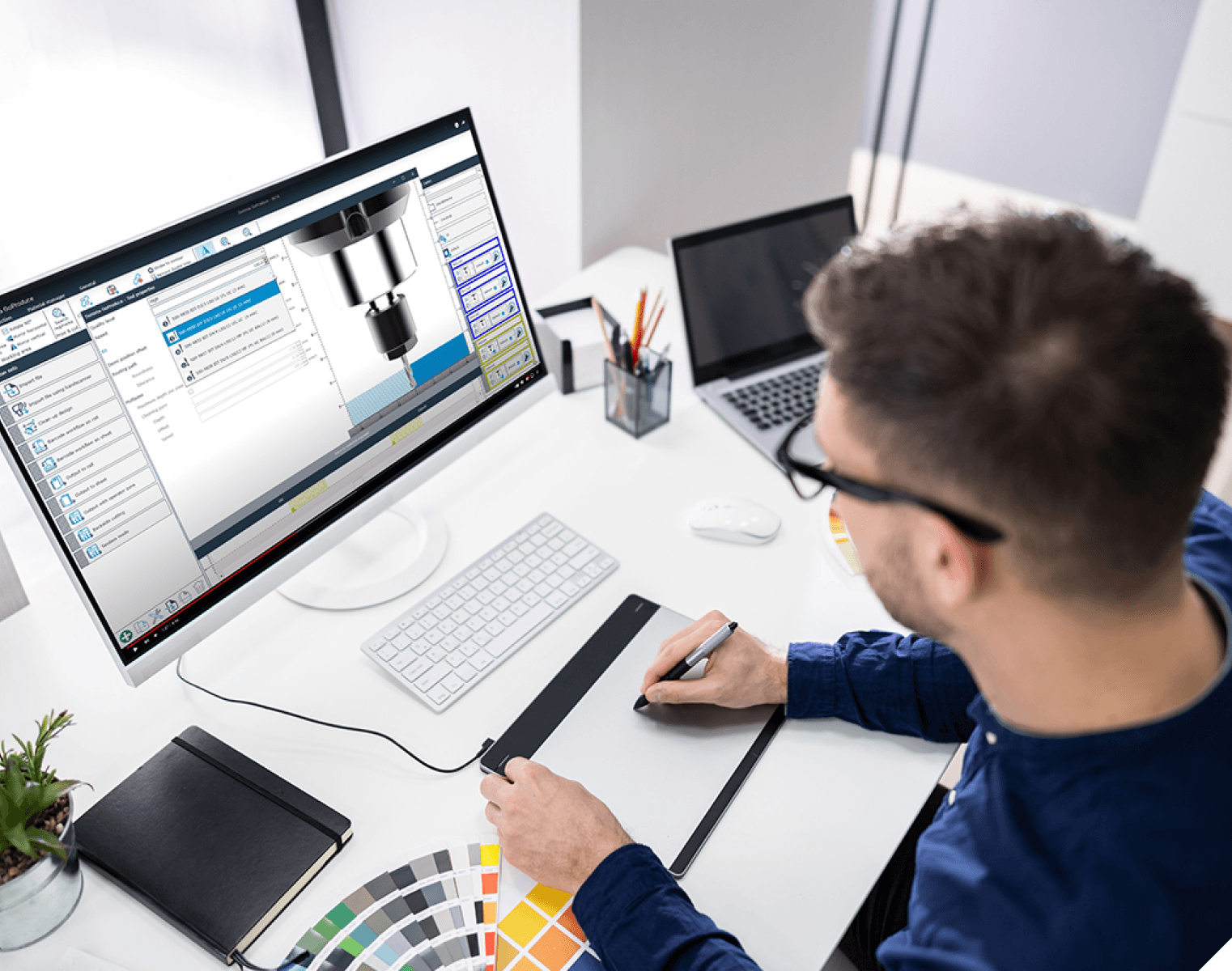 A person designs a 3D model on a computer using a graphic tablet at a desk with a laptop, color swatches, and office supplies.