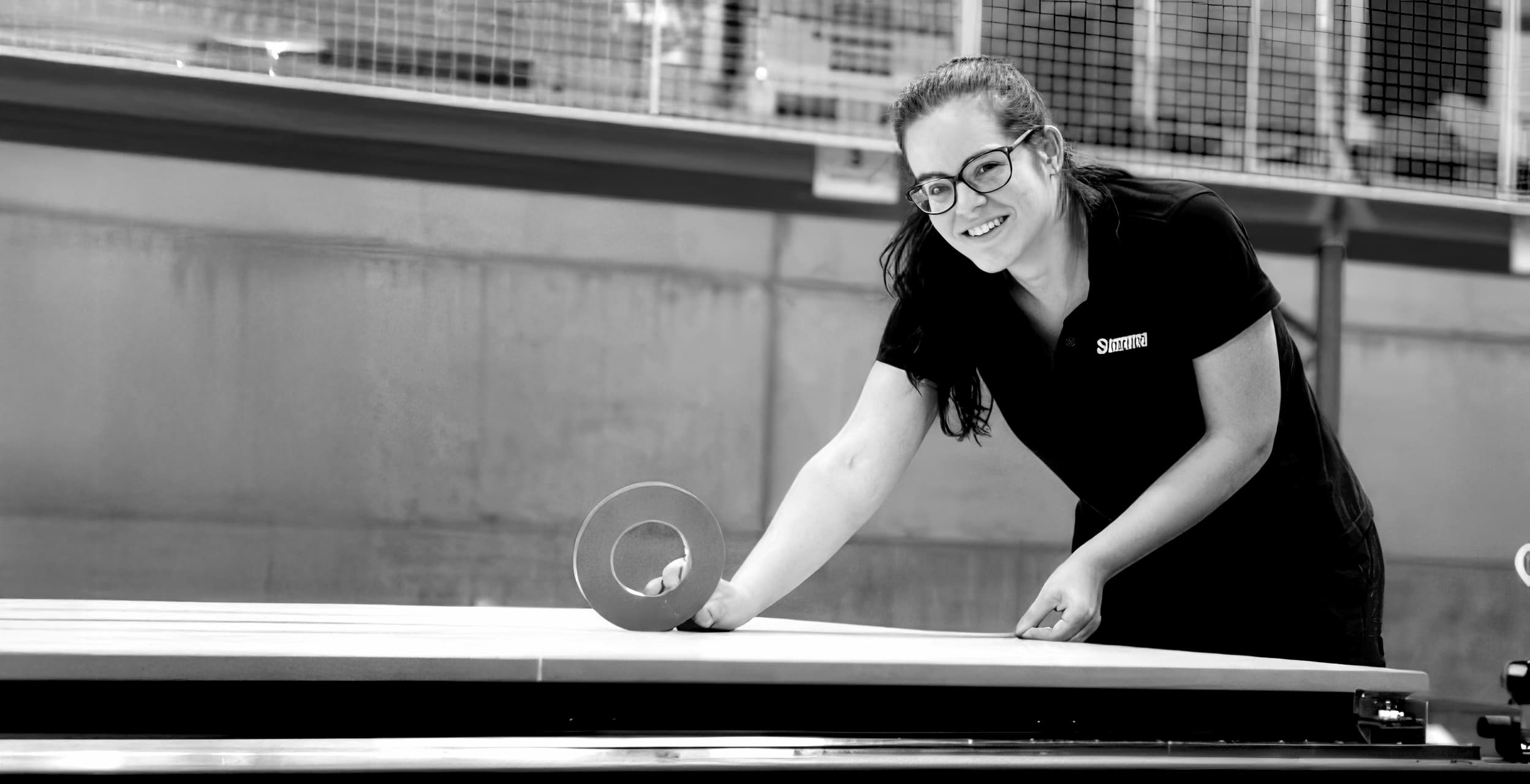 Woman in glasses smiling, applying tape to a large flat surface in an industrial setting. Black and white photo.