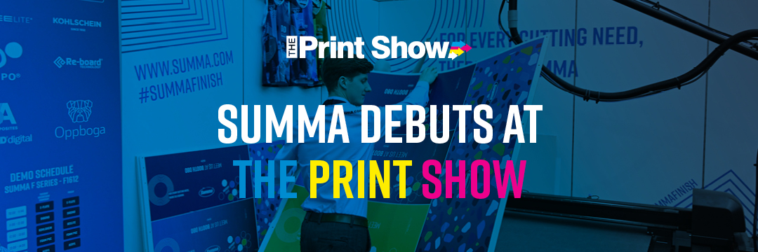 At The Print Show exhibition, a Summa Sales Support team member places a printed cardboard sheet onto a Summa flatbed cutter, preparing it to cut it into boxes. The Summa booth is visible in the background, with the text 'Summa debuts at The Print Show' alongside The Print Show logo overlaying the image.